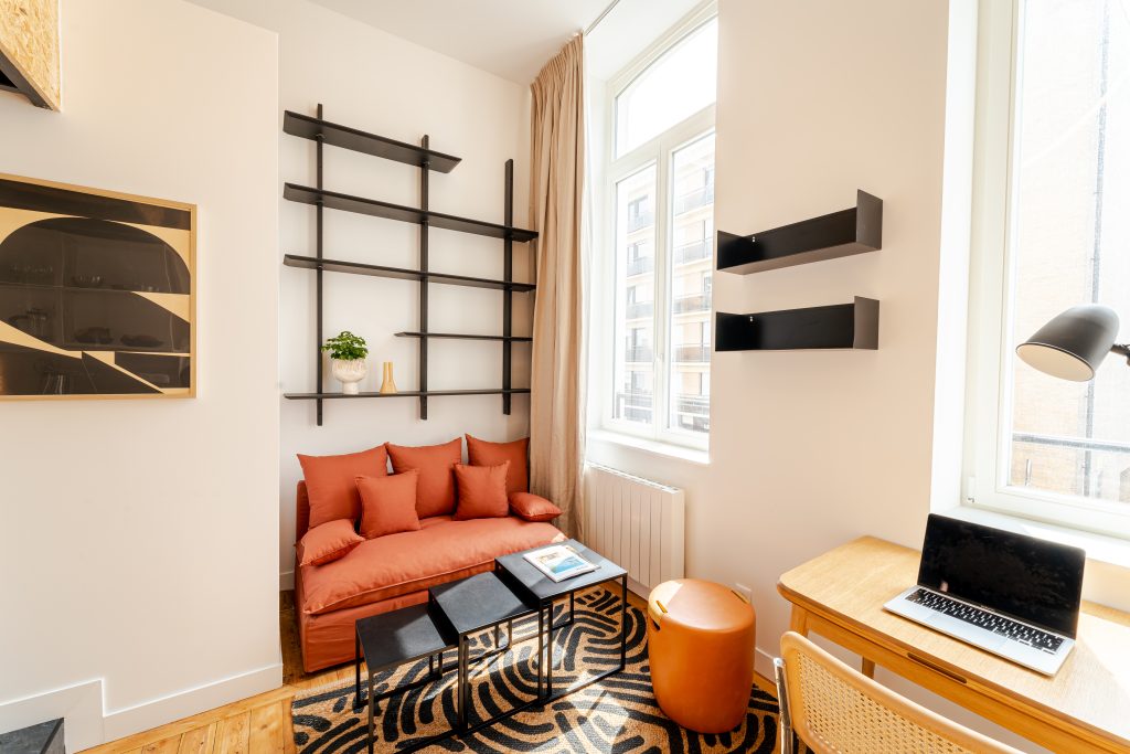 Studio meublé avec canapé orange, étagères et bureau près de la fenêtre