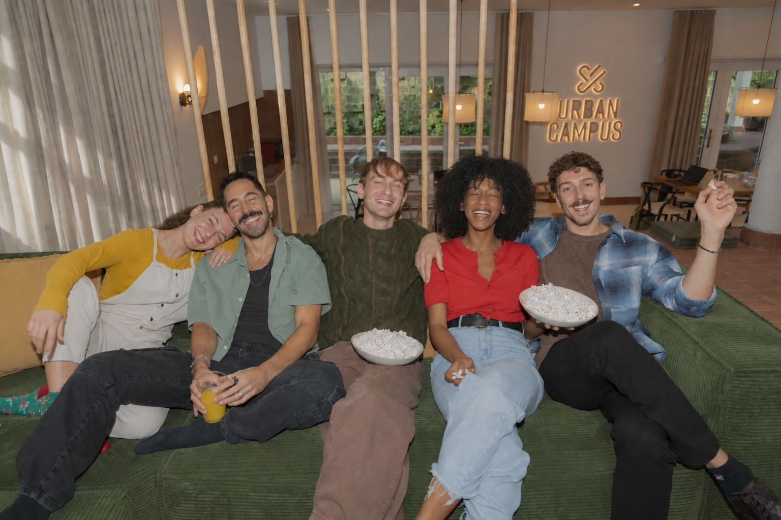 Cinq résidents souriants partagent du pop-corn sur un canapé vert dans un salon coliving lumineux Urban Campus
